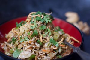 A close-up of a bowl filled with Oriental Salad, featuring shredded cabbage, carrots, crispy noodles, green onions, and chopped cilantro, with a fork to the side.