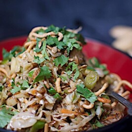 A close-up of an Oriental Salad with shredded cabbage, sliced grapes, crunchy noodles, and fresh cilantro, served in a red and black bowl.
