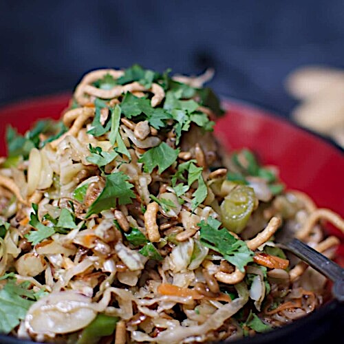 A close-up of an Oriental Salad with shredded cabbage, sliced grapes, crunchy noodles, and fresh cilantro, served in a red and black bowl.