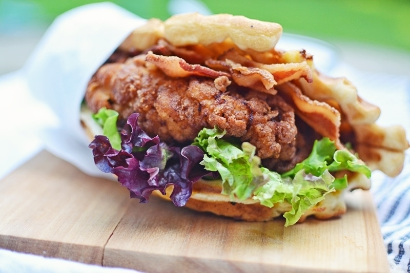 A close-up of a Crispy Chicken Waffle Wrap featuring fried chicken, crispy bacon, fresh lettuce, and a waffle bun, wrapped partially in paper and placed on a wooden board.