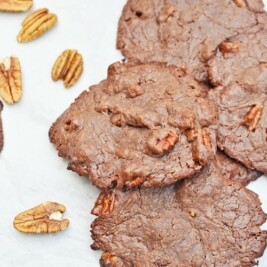 Six chewy chocolate cookies with pecans are scattered on a white surface, with several whole pecan halves placed nearby. The cookies have a rough, rustic texture and visible pecan pieces inside.