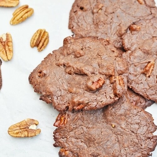 Six chewy chocolate cookies with pecans are scattered on a white surface, with several whole pecan halves placed nearby. The cookies have a rough, rustic texture and visible pecan pieces inside.