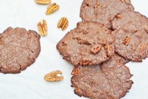 Several chewy chocolate cookies with visible pieces of pecan nuts are scattered on a white surface, with a few whole pecan halves lying nearby.