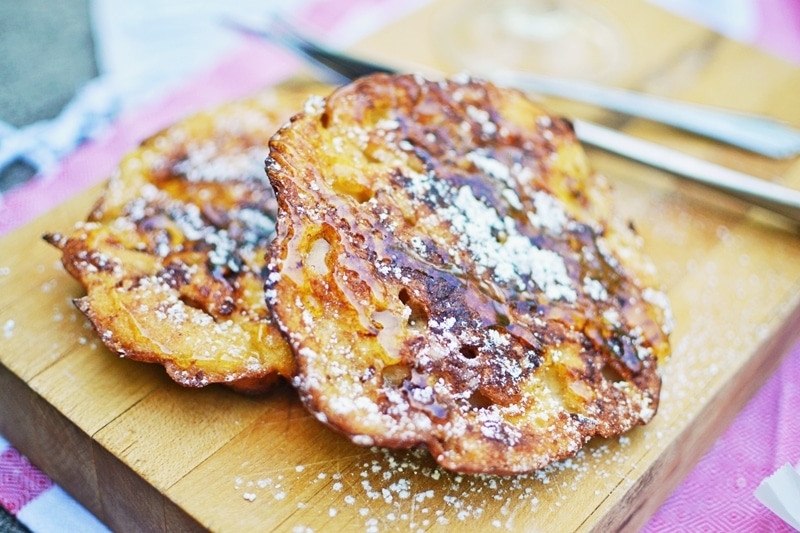 Two pieces of crispy, golden-brown French toast dusted with powdered sugar rest on a wooden board, with a fork and knife in the background.