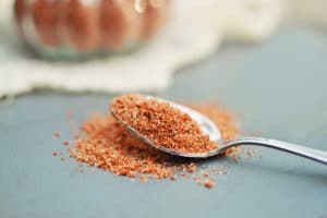 A close-up of a metal spoon holding a heap of reddish-brown spice rub, with some mix spilled onto a gray surface. A blurred jar appears in the background.