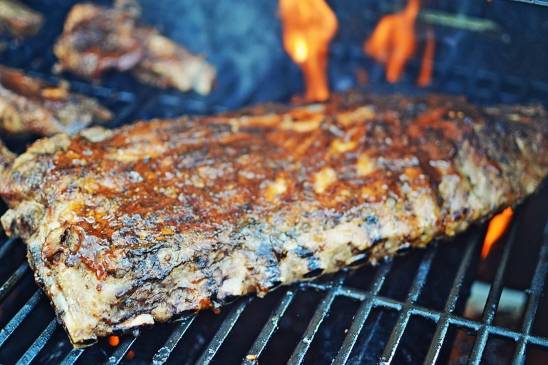 A rack of seasoned ribs is grilling on a barbecue with visible flames in the background, creating a smoky, charred texture on the meat glazed with tangy key lime BBQ sauce.