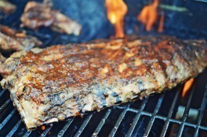 A rack of ribs with a browned, seasoned crust cooks on a grill over open flames, glazed with tangy key lime BBQ sauce, against a smoky background.