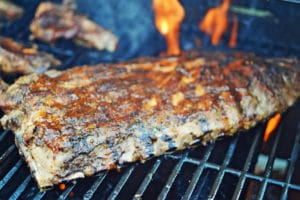 A rack of seasoned ribs is grilling on a barbecue with visible flames in the background, creating a smoky, charred texture on the meat glazed with tangy key lime BBQ sauce.