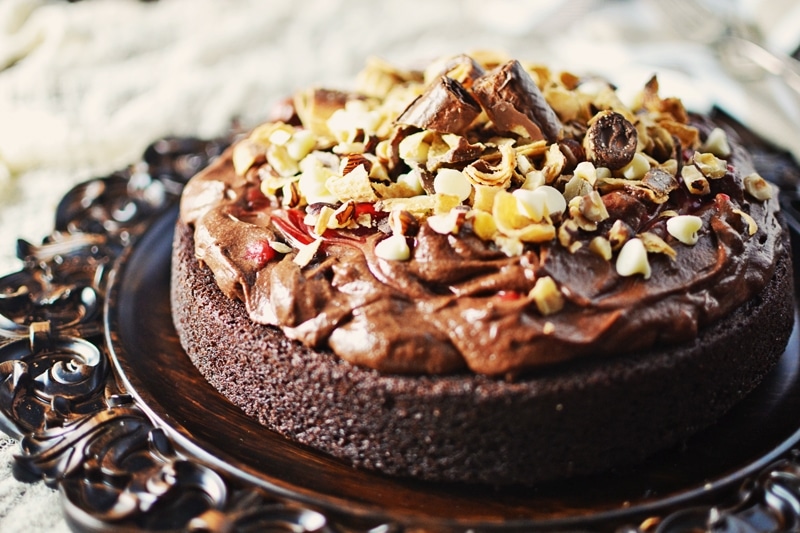 A rich Chocolate Mousse Cake topped with creamy chocolate frosting, chopped nuts, and chunks of chocolate, displayed on an ornate dark platter.