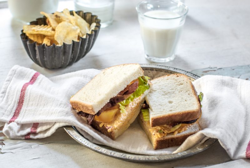 A black metal bowl with potato chip, glass of milk, and a sliced loaded BLT sandwich with Pepperidge Farm hearty white bread.