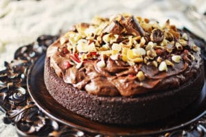 A rich Chocolate Mousse Cake topped with creamy chocolate frosting, chopped nuts, and chunks of chocolate, displayed on an ornate dark platter.