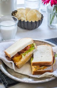 A plate with a Loaded BLT Sandwich cut in half on white bread, packed with bacon, lettuce, and tomato, sits on a napkin. In the background are a dish of potato chips, a glass of milk, and a vase with flowers.