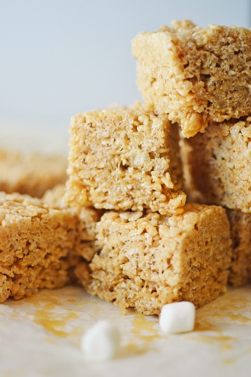 A close-up of stacked Rice Crispy Treats with a golden brown color, sitting on parchment paper. Two small white marshmallows are in the foreground.