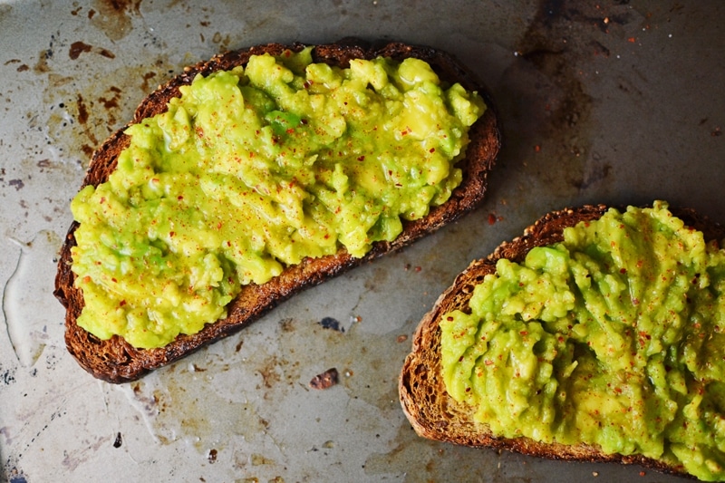 Two slices of toasted bread topped with creamy avocado mash and sprinkled with seasoning create a classic Avocado Toast, served on a metal baking sheet.