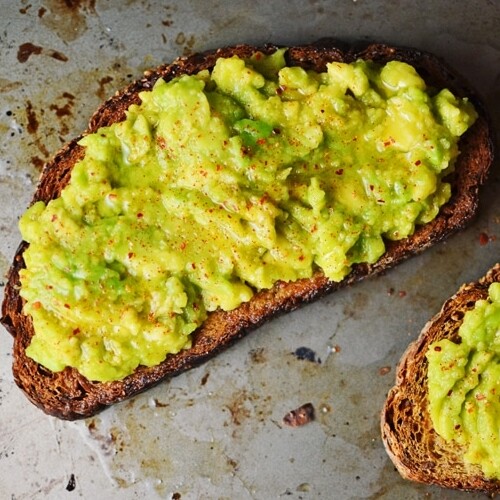 Two slices of toasted bread topped with creamy mashed avocado and sprinkled with red pepper flakes create a classic Avocado Toast, served on a metal surface.
