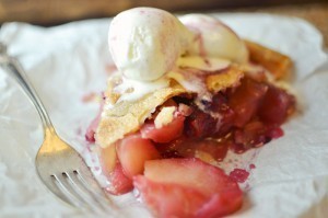 A slice of Mulled Apple and Blackberry Pie with a flaky crust is served on white parchment paper, topped with two scoops of vanilla ice cream, next to a silver fork.