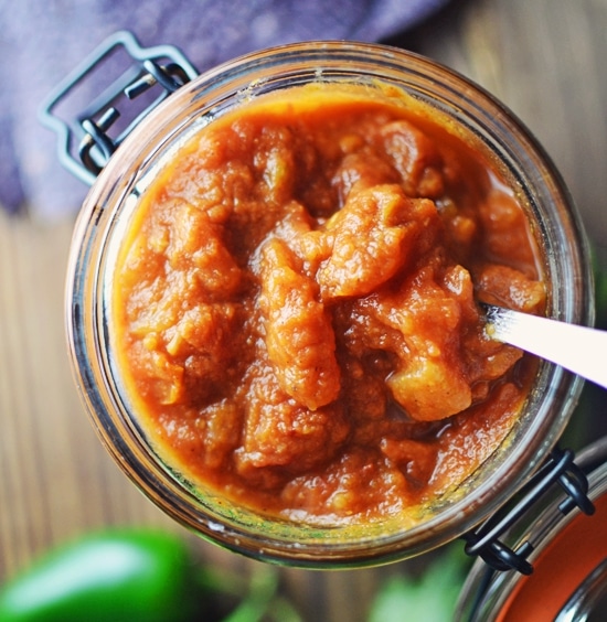 An overhead view of a glass jar filled with chunky Pumpkin Salsa, with a metal spoon inside. The thick, textured salsa sits in the open jar on a wooden surface.
