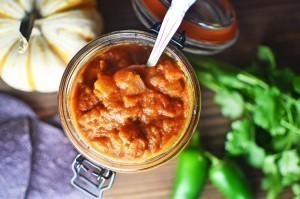 A jar of Pumpkin Salsa with a spoon in it sits on a wooden table, next to a small pumpkin, fresh cilantro, and green jalapeños.