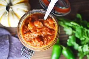 A jar of chunky red pumpkin salsa with a spoon inside, surrounded by a pumpkin, fresh cilantro, and green jalapeño peppers on a wooden surface.