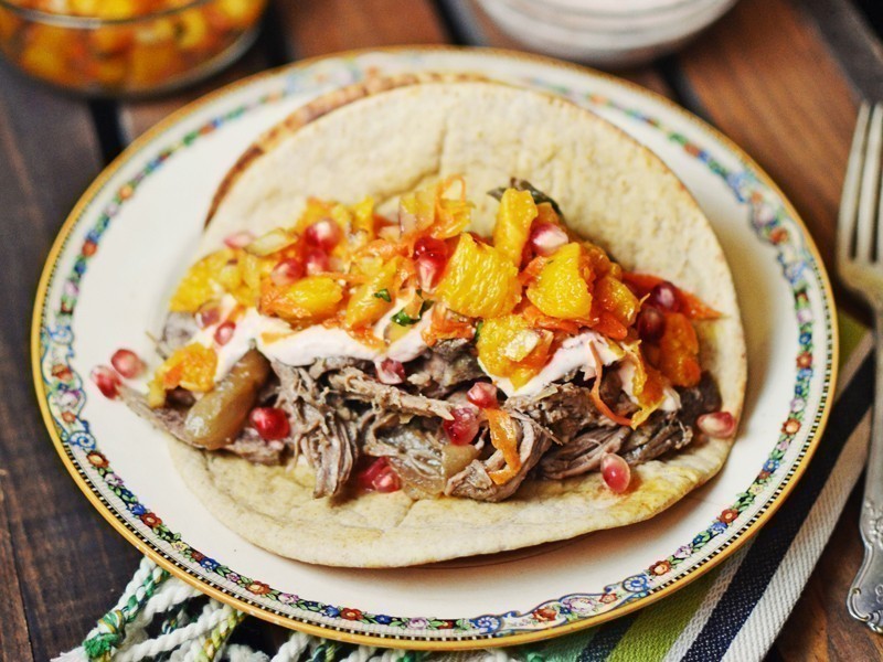 A plate with Shredded Lamb Tacos with Orange Salsa—topped with juicy shredded meat, orange chunks, pomegranate seeds, and creamy sauce—sits on a colorful patterned plate on a wooden table.