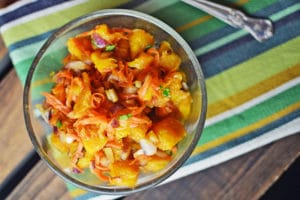 A glass bowl filled with a colorful salad of chopped mango, shredded carrots, red onion, and herbs sits on a striped green, blue, yellow, and white cloth beside a metal fork—perfect alongside Shredded Lamb Tacos with Orange Salsa.