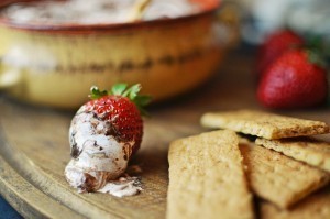 A chocolate-dipped strawberry with cream rests on a wooden board beside several rectangular crackers, perfect for dipping into a rich Smores Dip. A blurred bowl and whole strawberries are in the background.