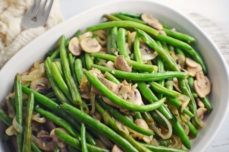 A white dish filled with Florida Snap Beans with Caramelized Onions and Mushrooms. In the background, a fork rests on a napkin, and a piece of bread or cauliflower is partially visible.