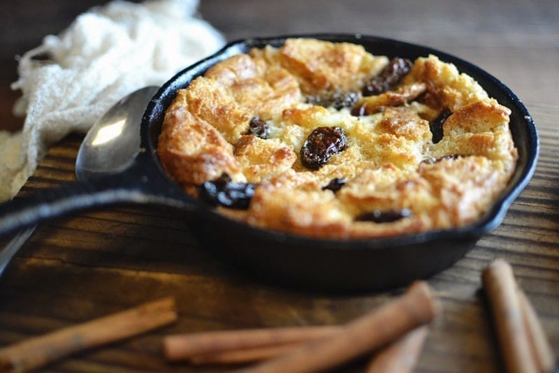 A classic bread pudding with raisins is baked in a black cast iron skillet, resting on a wooden surface with a spoon, white cloth, and cinnamon sticks nearby.