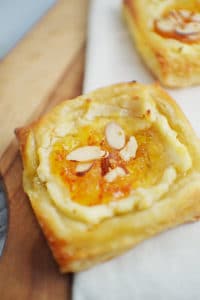 A close-up of a Marmalade Cream Cheese Danish pastry topped with creamy filling, orange marmalade, and sliced almonds, set on a wooden board with a white cloth.
