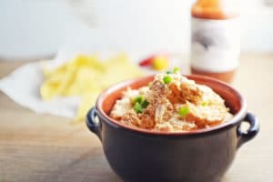 A black ceramic bowl filled with creamy chicken dip, topped with shredded meat and chopped green onions, sits on a wooden surface. In the background, tortilla chips and a bottle of hot sauce are softly blurred.