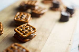 Chocolate Caramel Pretzel Bites are arranged on a wooden board, featuring square pretzels with chocolate and caramel sandwiched between them, while more pretzels and chocolate candies are blurred in the background.