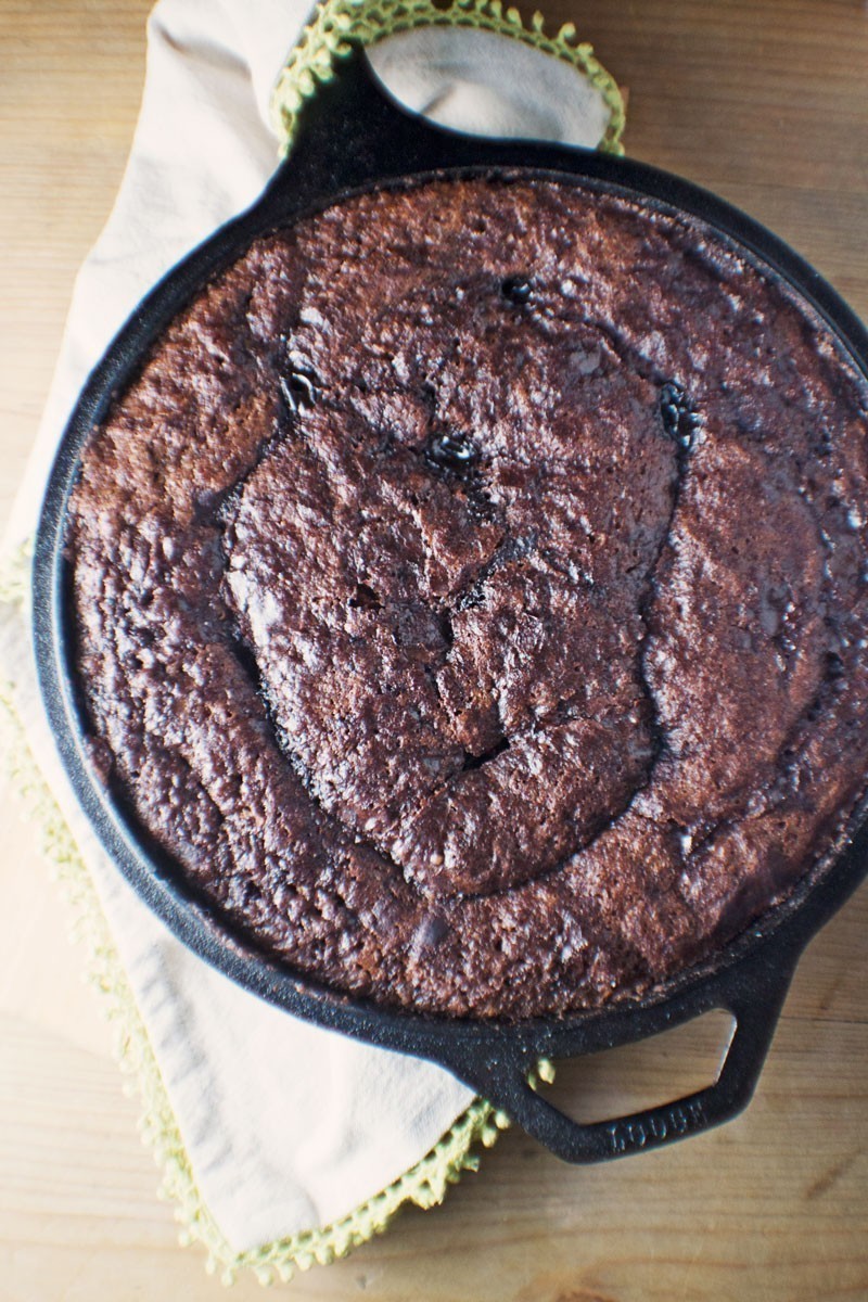 A freshly baked Mocha Fudge Cobbler in a black cast iron skillet rests on a cream-colored cloth with green trim, placed on a wooden surface.