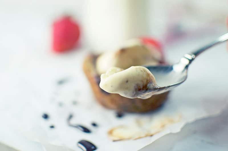 A close-up of a spoon holding a creamy dessert, reminiscent of sundae indulgence, with a pastry and chocolate sauce on parchment paper in the blurred background. A red berry is also visible.