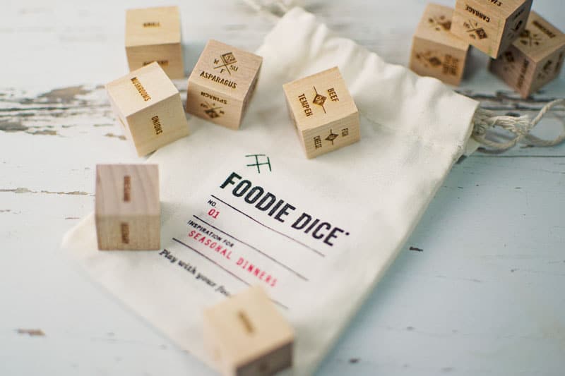 A cloth bag labeled FOODIE DICE lies on a blue wooden surface, surrounded by several wooden dice featuring various food ingredients and cooking methods—an inspiring gift idea for Mother’s Day.