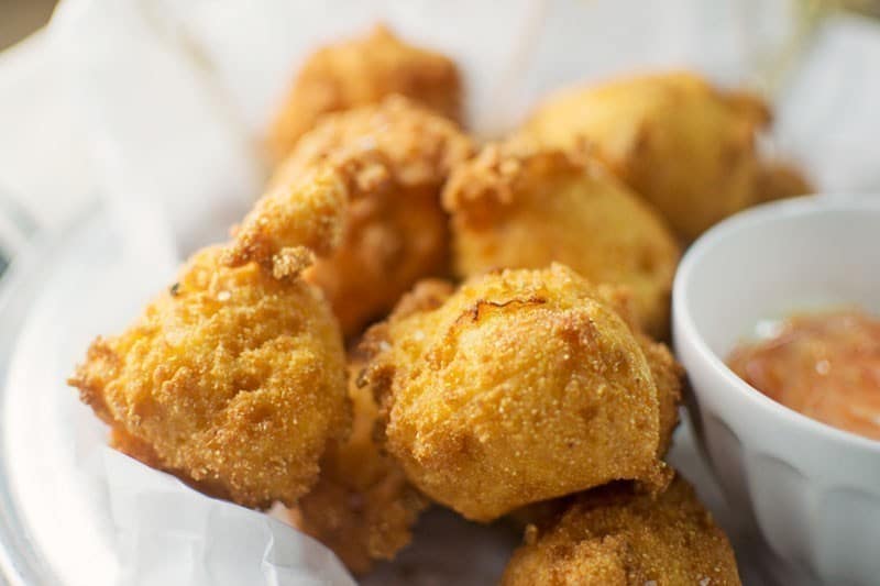 A close-up of golden-brown hushpuppies on a white plate lined with parchment paper, served with a small bowl of dipping sauce on the side.