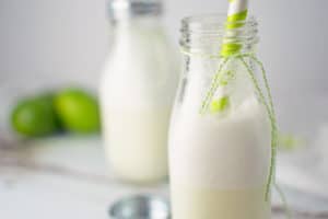 A glass milk bottle filled with frothy Brazilian Lemonade, topped with a green striped straw and green string tied around the neck. Another similar bottle and fresh green limes are blurred in the background.
