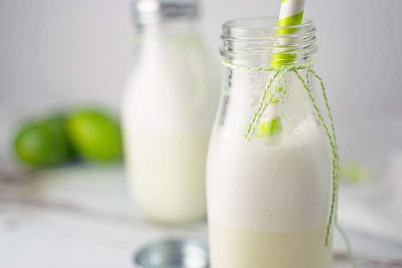A glass milk bottle filled with frothy Brazilian Lemonade, topped with a green striped straw and green string tied around the neck. Another similar bottle and fresh green limes are blurred in the background.