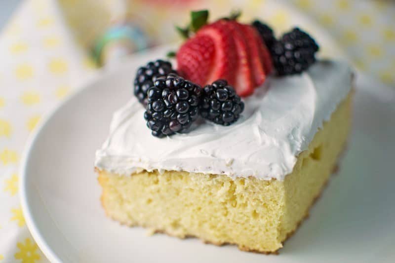 A slice of yellow cake with white frosting, inspired by a classic sheet cake recipe, topped with blackberries and a sliced strawberry, served on a white plate.