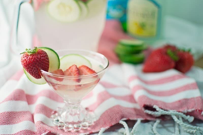 A refreshing cucumber drink, this glass of pink lemonade is garnished with fresh strawberries and cucumber slices, sitting on a pink-and-white striped cloth, with more strawberries and a pitcher in the background.