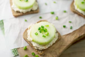 A slice of bread topped with a creamy spread, a thick slice of cucumber crudites, and chopped green onions, placed on parchment paper with extra green onion pieces scattered around.