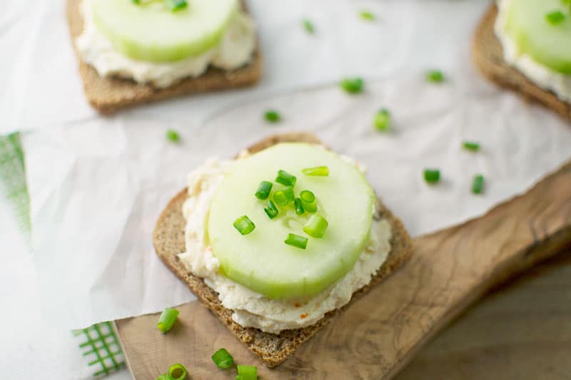 A slice of bread topped with a creamy spread, a thick slice of cucumber crudites, and chopped green onions, placed on parchment paper with extra green onion pieces scattered around.