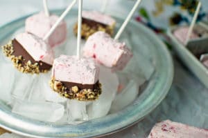 Fruity Ice Pops in pink frozen yogurt with chocolate and crushed nuts on the bottom sit on a silver plate of ice cubes. Each pop is on a stick, with a colorful cloth adding charm in the background.
