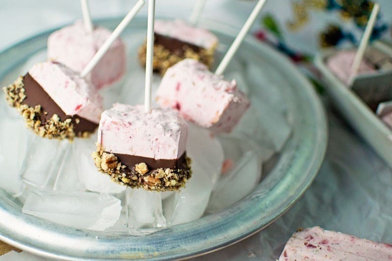 Fruity Ice Pops in pink frozen yogurt with chocolate and crushed nuts on the bottom sit on a silver plate of ice cubes. Each pop is on a stick, with a colorful cloth adding charm in the background.