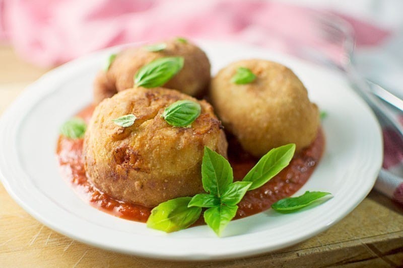 Three golden-brown arancini balls sit on a white plate over a bed of marinara sauce, garnished with fresh basil leaves—these savory bites are reminiscent of Pizza Bombs. A fork and a pink napkin are visible in the background.