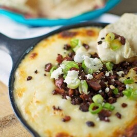 A small cast iron skillet filled with melted Chorizo Queso Dip, topped with chopped green onions, bacon bits, and crumbled cheese. A tortilla chip is dipped into the cheesy mix, with more chips in a dish in the background.