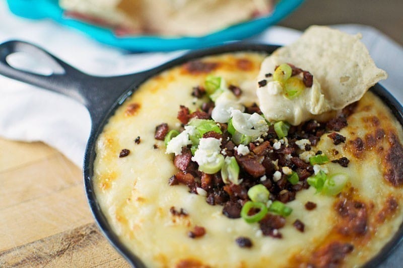 A small cast iron skillet filled with melted Chorizo Queso Dip, topped with chopped green onions, bacon bits, and crumbled cheese. A tortilla chip is dipped into the cheesy mix, with more chips in a dish in the background.