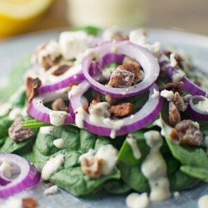 A fresh Spinach Salad with Feta Dressing, topped with red onion rings, crumbled bacon, and creamy feta cheese, served on a plate with lemons in the background.