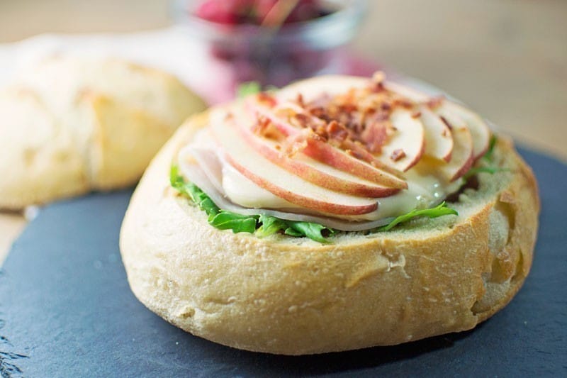 A round bread bowl filled with leafy greens, sliced turkey, melted cheese, thin apple slices, and crumbled bacon sits on a dark surface—reminiscent of a Triple Meat with Brie Picnic Sandwich—with another bread bowl and cherries in the blurred background.