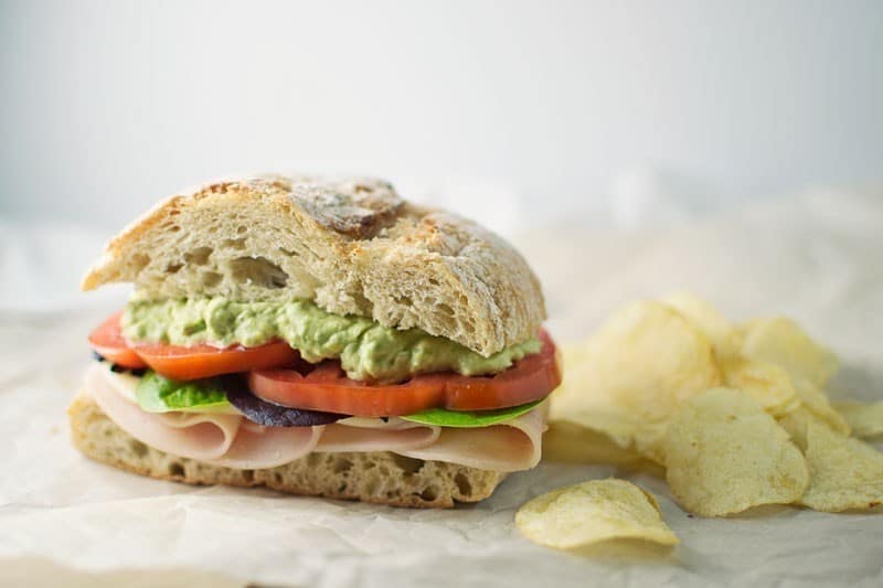 A Roast Turkey with Garlic Avocado Mayo Sandwich featuring turkey, lettuce, tomato, and creamy garlic avocado spread on crusty bread sits on parchment paper beside a small pile of potato chips.