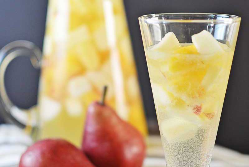 A glass of Autumn Sangria with chopped fruit, including pears and pineapple, sits in front of a matching pitcher. Two whole red pears rest on a white and gray striped cloth nearby.